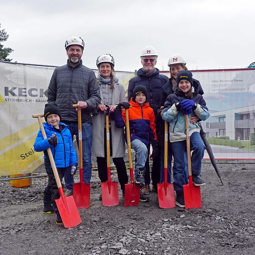 Eine Gruppe von Erwachsenen und Kindern mit Schaufeln steht an einer Baustelle. Sie tragen Helme und Winterkleidung. Ein großes Banner ist hinter ihnen sichtbar.
