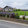 Eine Blaskapelle in traditionellen Uniformen spielt Instrumente, während sie eine Straße entlanggehen, mit einem Haus und einem Grasfeld im Hintergrund.