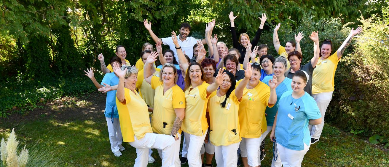 Eine Gruppe von Gesundheitspersonal in gelben und blauen Uniformen posiert im Freien für ein Foto, mit Bäumen im Hintergrund.