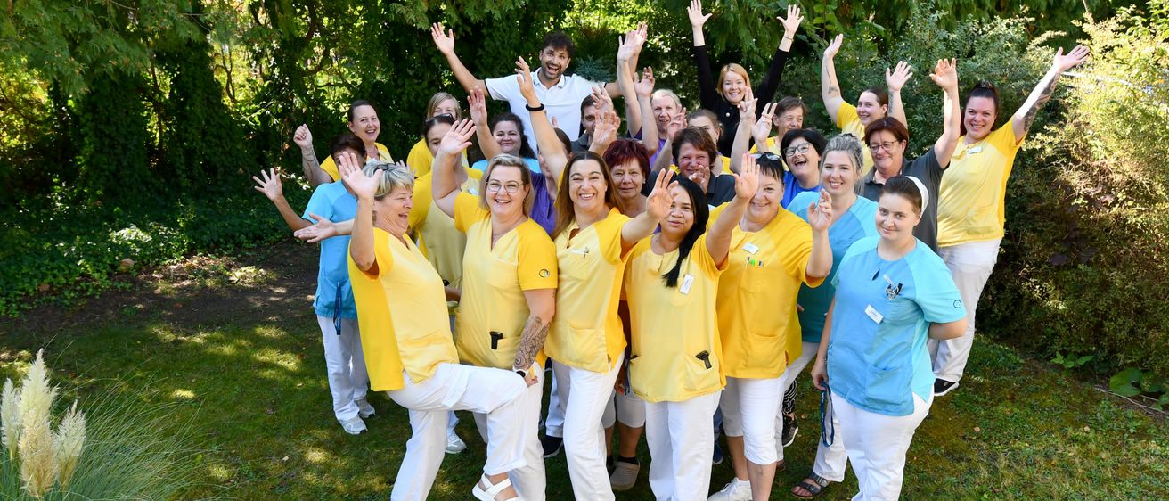 Eine Gruppe von Gesundheitspersonal in gelben und blauen Uniformen posiert im Freien für ein Foto, mit Bäumen im Hintergrund.