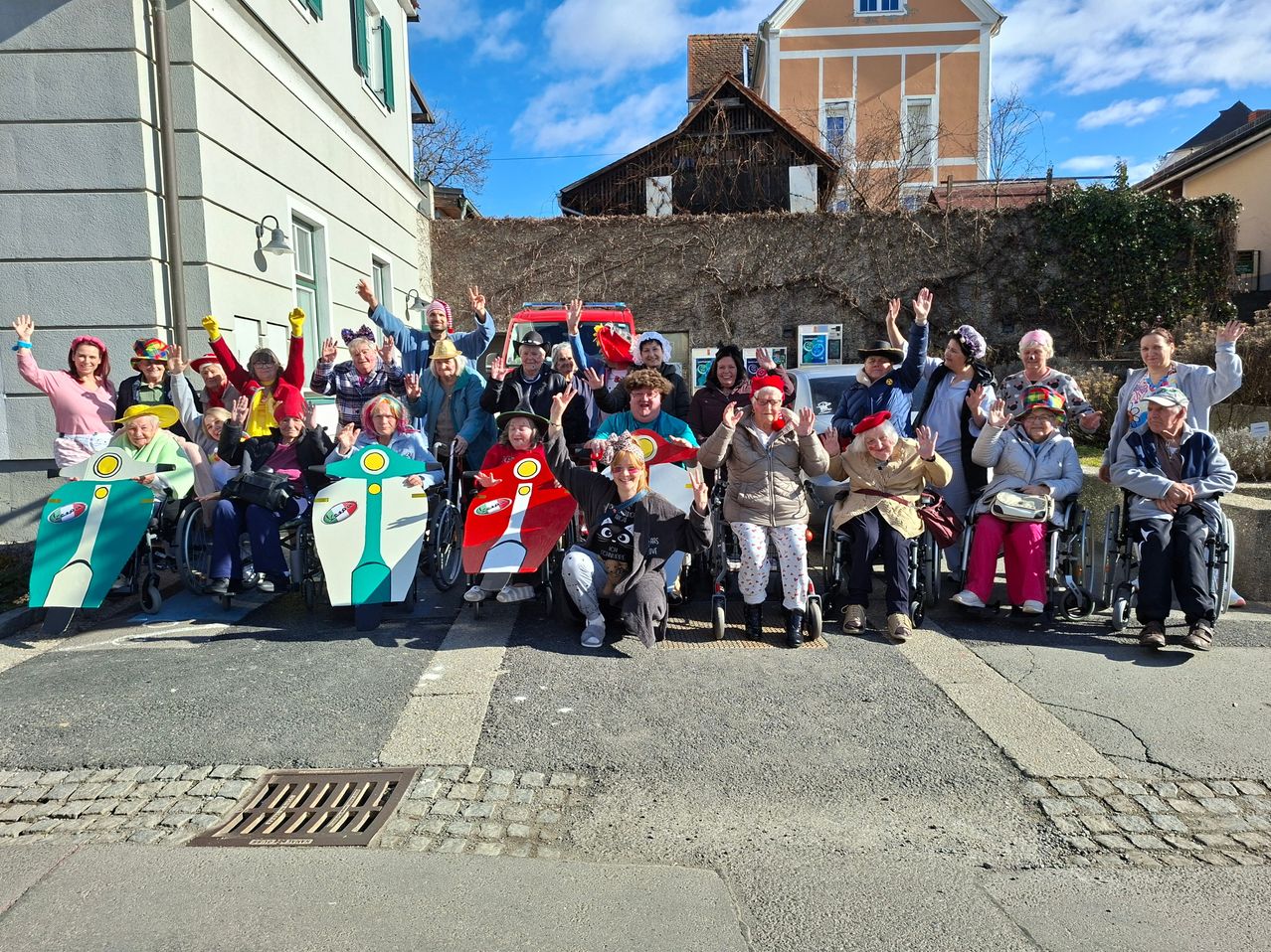 Eine Gruppe älterer Menschen in Rollstühlen posiert für ein Foto, einige tragen Hüte und Handschuhe. Sie haben bunte Schilder auf dem Rücken. Dahinter steht eine Mauer mit Kletterpflanzen.