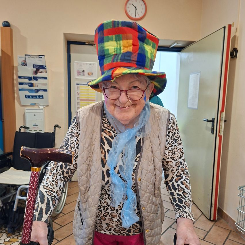 Eine ältere Frau mit einem farbenfrohen Hut, Leopardenmusterkleidung und einer Brille steht lächelnd in einem Flur und hält einen Stock. Hinter ihr steht ein Rollstuhl, eine Uhr und eine offene Tür.