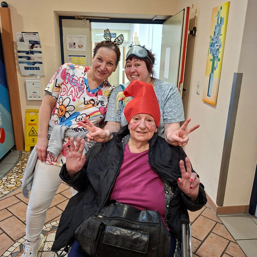 Drei Frauen posieren für ein Foto, von denen eine älter ist und in einem Rollstuhl sitzt. Sie alle lächeln und machen Friedenszeichen.