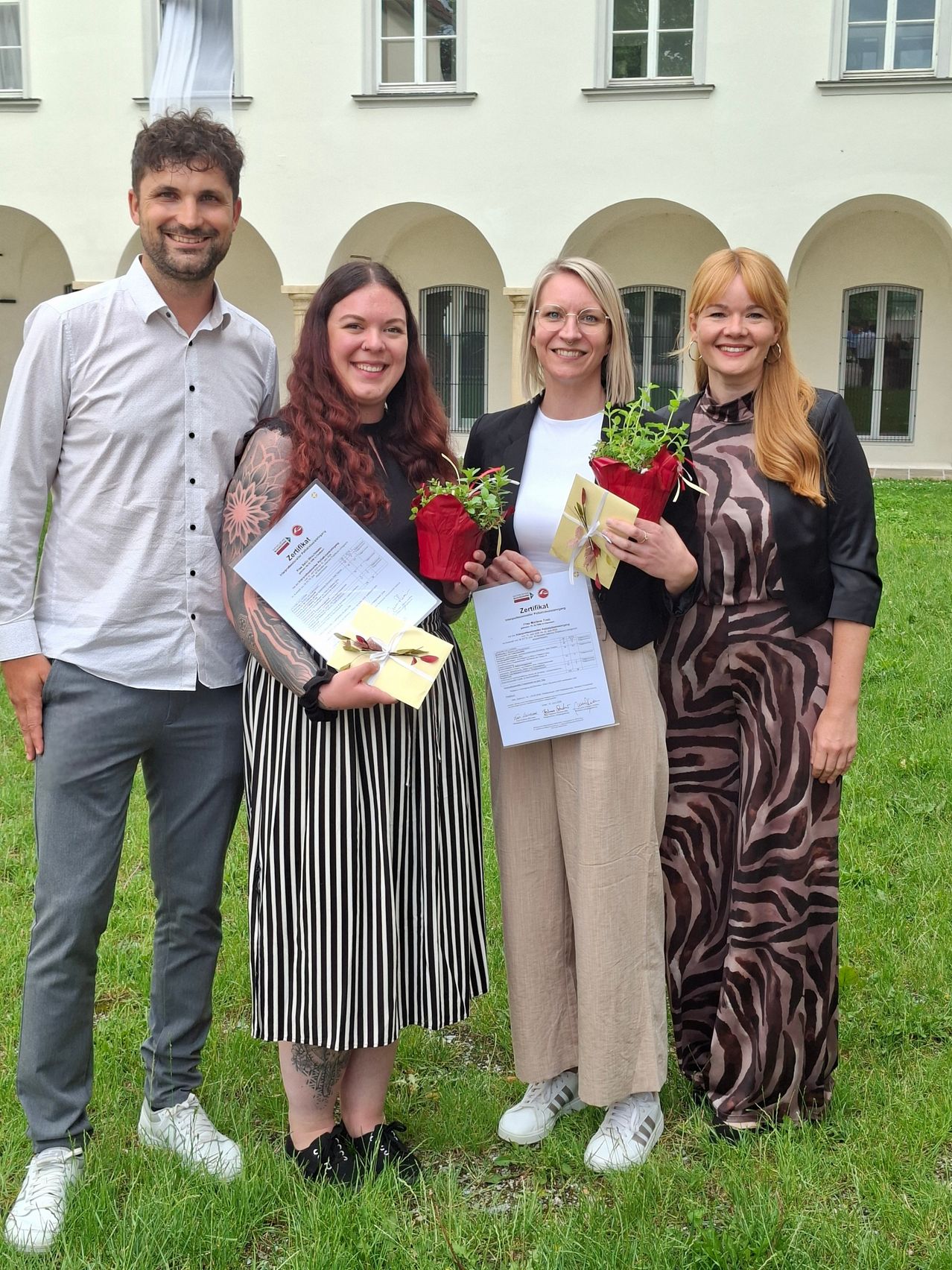 Vier Personen stehen lächelnd auf einem Rasen und halten Urkunden und Blumen. Ein Mann in einem weißen Hemd und Jeans steht links. Die anderen sind Frauen, zwei in Schwarz und Weiß, eine in einem Zebra-Druck-Kleid. Sie halten alle Urkunden und kleine Topfpflanzen.