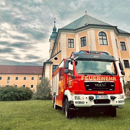 Ein roter Feuerwehrwagen mit der Nummer 4311 ist vor einem großen Gebäude geparkt. Das Gebäude hat ein kuppelförmiges Dach und viele Fenster. Vor dem Gebäude stehen Pflanzen und Büsche.