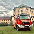 Ein roter Feuerwehrwagen mit der Nummer 4311 ist vor einem großen Gebäude geparkt. Das Gebäude hat ein kuppelförmiges Dach und viele Fenster. Vor dem Gebäude stehen Pflanzen und Büsche.