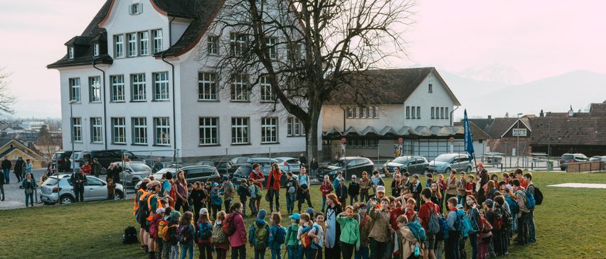 A group of children with backpacks are gathered in a circle in a grassy field in front of a white building with many windows and a bare tree.