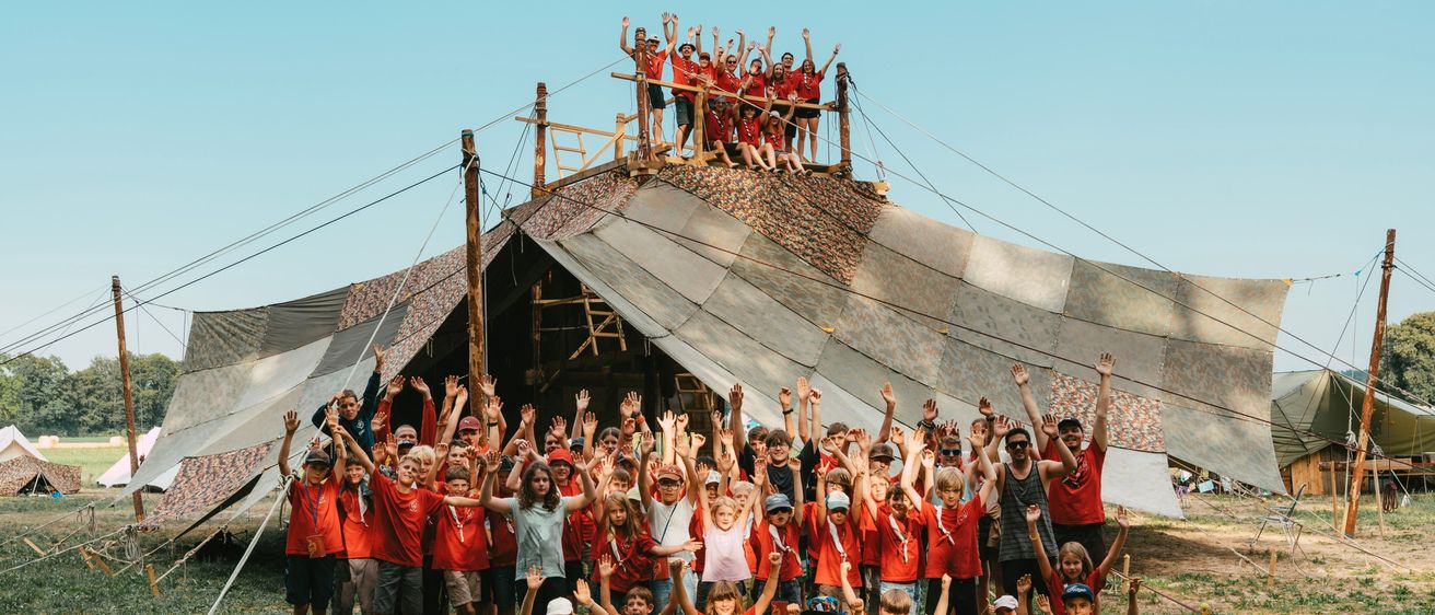 Eine große Gruppe von Kindern und Erwachsenen in roten T-Shirts versammelt sich auf einem Grasfeld und posiert für ein Foto vor einem großen Zelt.