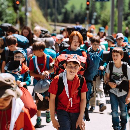 Eine Gruppe von Kindern in roten und blauen Uniformen und Mützen, mit Rucksäcken, läuft eine Straße entlang. Sie sind wahrscheinlich auf einem Schulausflug.