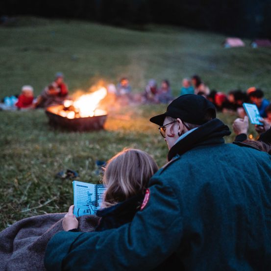 Eine Familie sitzt nachts um ein Lagerfeuer herum, während andere zusehen. Ein Erwachsener liest einem Kind ein Buch vor.
