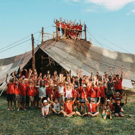 Eine Gruppe von Kindern in roten T-Shirts posiert vor einem Zelt unter blauem Himmel, einige stehen auf dem Zeltdach.