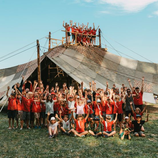 Eine Gruppe von Kindern in roten T-Shirts posiert vor einem Zelt unter blauem Himmel, einige stehen auf dem Zeltdach.