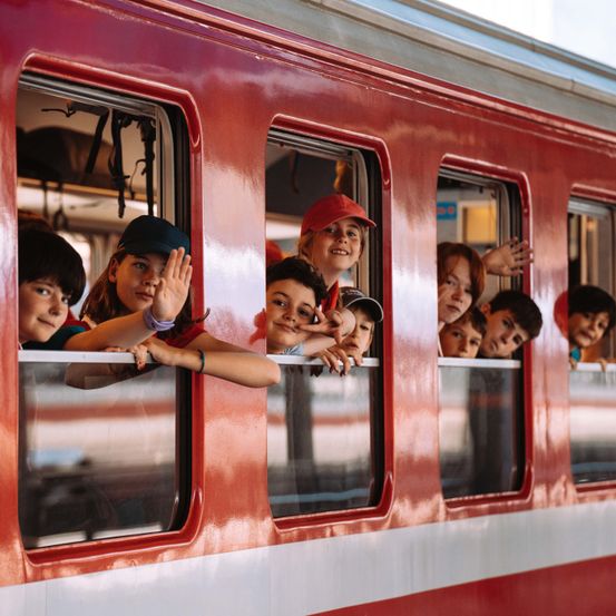 Eine Gruppe von Kindern mit aufgeregten Gesichtern schaut aus den Fenstern eines roten Zuges. Einige tragen Hüte und ein Mädchen winkt.