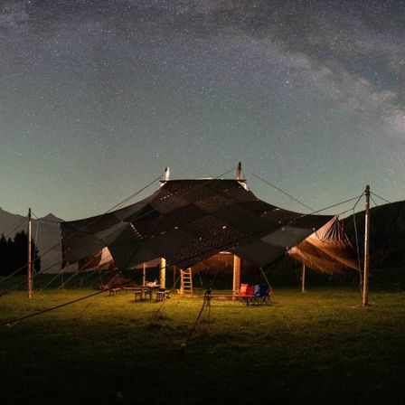 Ein großes Zelt steht auf einer Wiese unter einem sternenklaren Nachthimmel. In der Nähe befinden sich Stühle, ein Tisch und eine Leiter. Der Himmel ist mit Sternen und einem hellen Mond gefüllt.