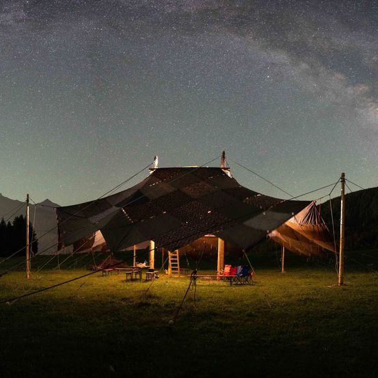 Ein großes Zelt steht auf einer Wiese unter einem sternenklaren Nachthimmel. In der Nähe befinden sich Stühle, ein Tisch und eine Leiter. Der Himmel ist mit Sternen und einem hellen Mond gefüllt.