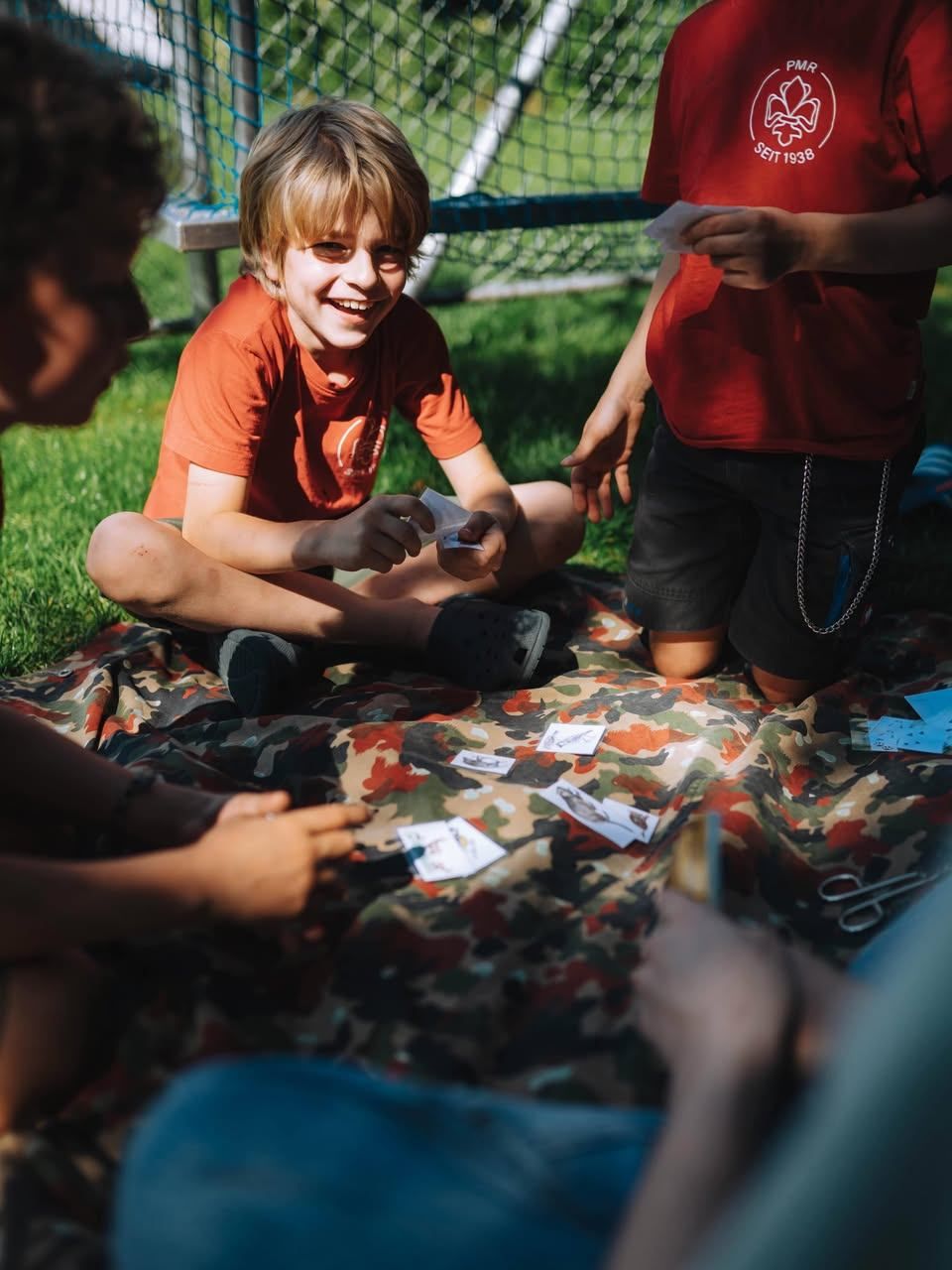 A group of children are sitting on a camouflage blanket on the grass, playing a card game. One child is smiling and holding a card.