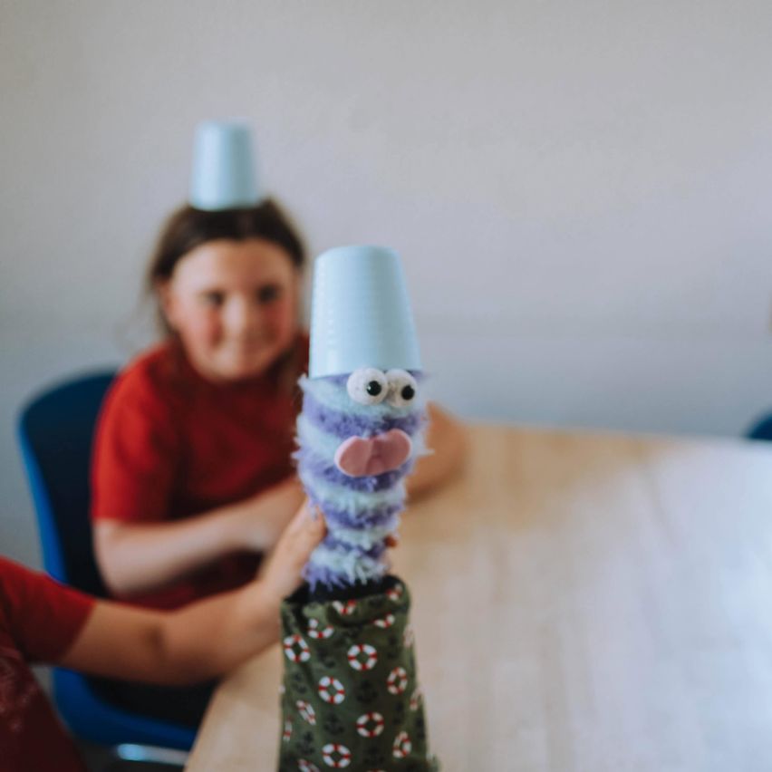 Two children are playing a game with blue cups and a purple sock puppet. The girl in the red shirt is watching.