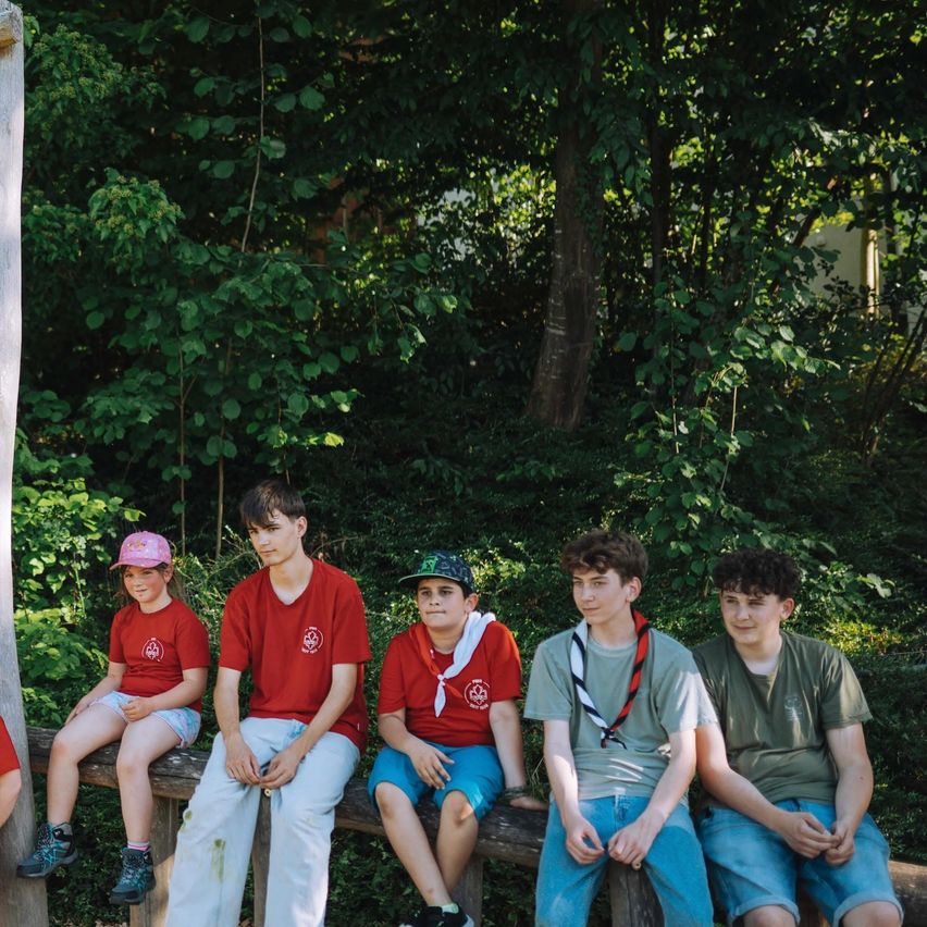 Five young boys and girls are sitting on a wooden bench in a forest, wearing matching red shirts and hats. They are smiling and posing for a photo.