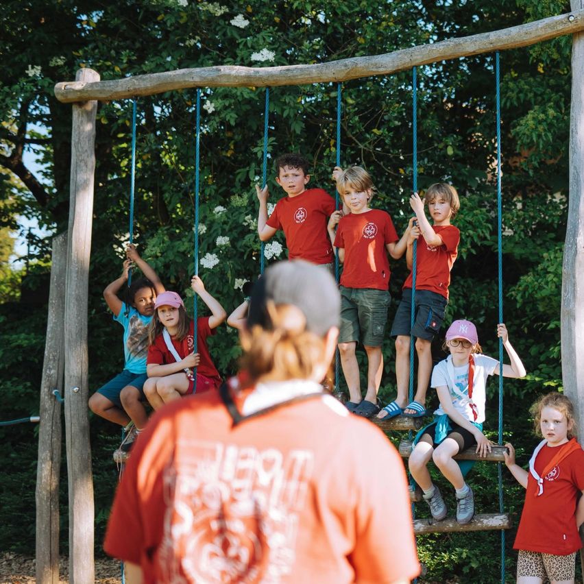 A group of children in red shirts are playing on swings in a forest setting, supervised by an adult in an orange shirt.