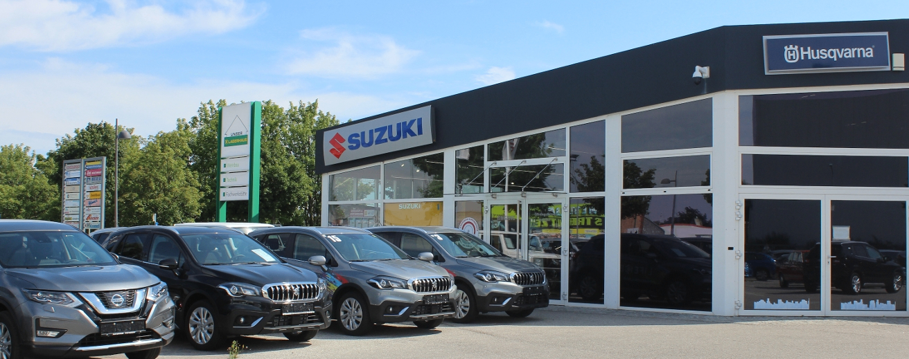 Ein Suzuki-Autohaus mit mehreren Autos vor dem Gebäude geparkt an einem sonnigen Tag. Das Gebäude hat Glastüren und -fenster und ein Schild mit dem Wort Suzuki.