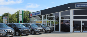 Ein Suzuki-Autohaus mit mehreren Autos vor dem Gebäude geparkt an einem sonnigen Tag. Das Gebäude hat Glastüren und -fenster und ein Schild mit dem Wort Suzuki.