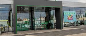 Ein modernes Gebäude mit einem Schild 'UNSER X LAGERHAUS' über dem Eingang. Der Eingang hat automatische Schiebetüren. Ein großes Fenster zeigt das Innere. Ein Banner mit einer Frau auf einem Schwimmkörper befindet sich rechts.