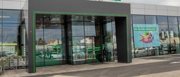 Ein modernes Gebäude mit einem Schild 'UNSER X LAGERHAUS' über dem Eingang. Der Eingang hat automatische Schiebetüren. Ein großes Fenster zeigt das Innere. Ein Banner mit einer Frau auf einem Schwimmkörper befindet sich rechts.