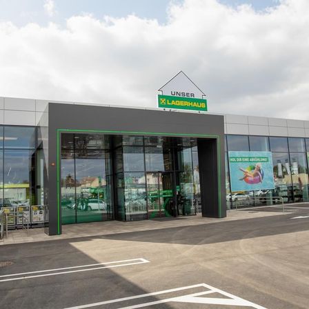 Außenansicht eines Lagerhauses mit dem Schild 'UNSER X LAGERHAUS'. Der Eingang hat Glastüren und grüne Verkleidungen. Ein Banner ist an der Seite des Gebäudes angebracht.