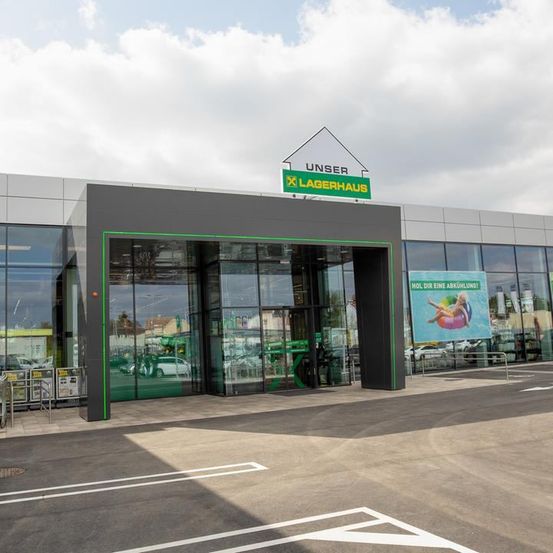 Außenansicht eines Lagerhauses mit dem Schild 'UNSER X LAGERHAUS'. Der Eingang hat Glastüren und grüne Verkleidungen. Ein Banner ist an der Seite des Gebäudes angebracht.
