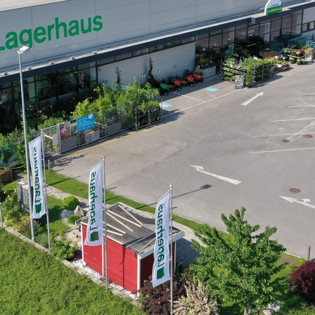 Luftaufnahme eines Lagerhaus-Geschäfts mit verschiedenen Pflanzen und Gartengeräten davor. Der Laden verfügt über einen großen Parkplatz mit Richtungspfeile.