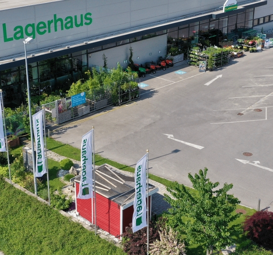 Luftaufnahme eines Lagerhaus-Geschäfts mit verschiedenen Pflanzen und Gartengeräten davor. Der Laden verfügt über einen großen Parkplatz mit Richtungspfeile.