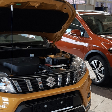 Ein orangefarbener SUV mit offenem Motorhaube in einer Ausstellung, der seinen Motor zeigt. Ein orangefarbenes Auto ist daneben geparkt.