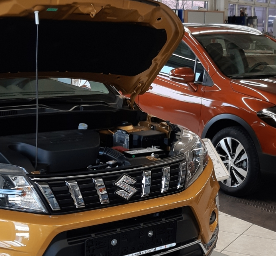 Ein orangefarbener SUV mit offenem Motorhaube in einer Ausstellung, der seinen Motor zeigt. Ein orangefarbenes Auto ist daneben geparkt.