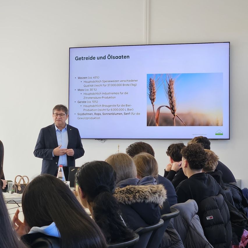 Ein Mann steht vor einer Präsentationsfolie und diskutiert über Getreide und Ölsaaten, wobei eine Folie verschiedene Feldfrüchte zeigt. Das Publikum, in Stühlen sitzend, hört aufmerksam zu.