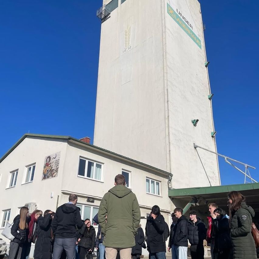 Eine Gruppe von Menschen steht vor einem hohen Gebäude mit einem grünen Dach und dem Wort 'UNSER' auf der Seite. Das Gebäude hat ein großes Fenster auf der linken Seite und eine Wandmalerei auf der rechten Seite.