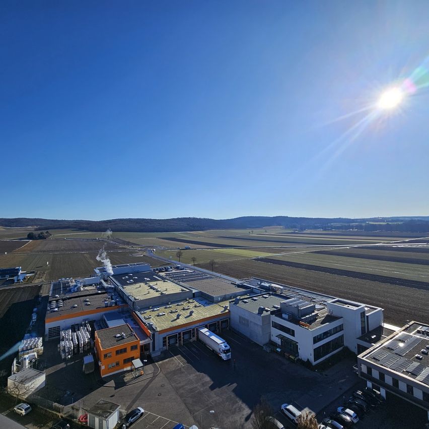 Eine Luftaufnahme eines großen Industriekomplexes mit mehreren Gebäuden und Fahrzeugen auf einem weiten offenen Feld unter einem strahlend blauen Himmel.