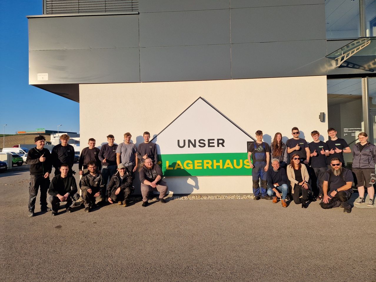 A group of people, dressed casually, are posing for a photo in front of a white building with a sign reading 'UNSER LAGERAUS'.