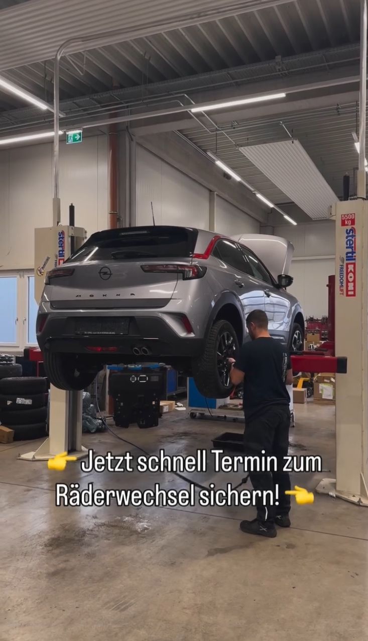 A man is working on a car lifted up in a garage. The car is silver with a black roof. There is a sign on the wall that reads 'Jetzt schnell Termin zum Räderwechsel sichern!'. There are other cars and tools in the garage.