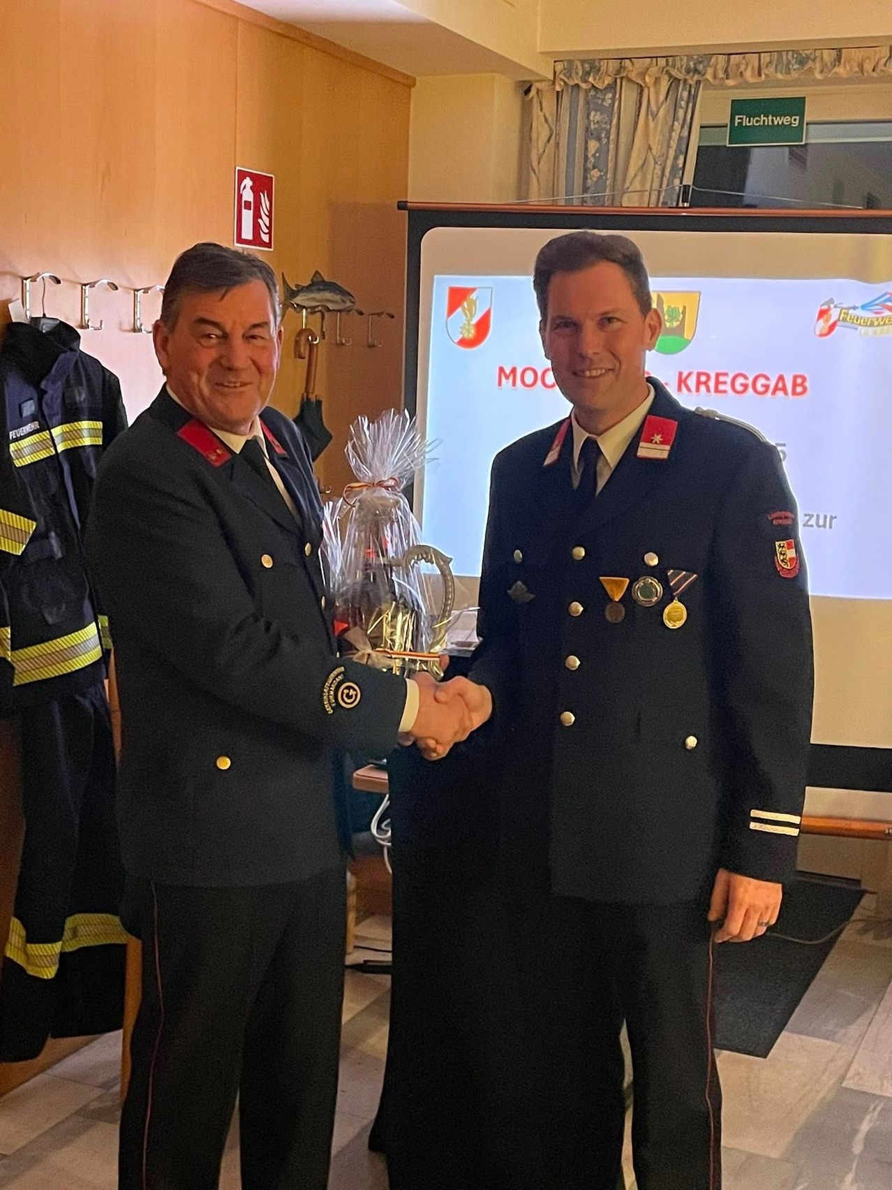 Zwei Männer in Militäruniformen tauschen einen Händedruck aus, während sie ein Geschenk halten. Hinter ihnen zeigt eine Projektorleinwand ein Logo. Ein Feuerwehrmannsuniform hängt an der Wand.