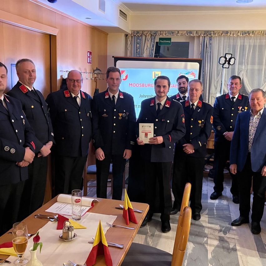 Mehrere Männer in Uniform stehen um einen Tisch mit einem Weinglas, einige halten eine Urkunde, mit einer Projektionsleinwand im Hintergrund.