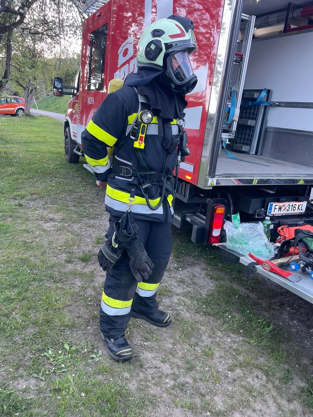Ein Feuerwehrmann in voller Montur, einschließlich Maske und Handschuhen, steht neben einem roten LKW mit einem Kennzeichen, das FW 316 KJ liest.