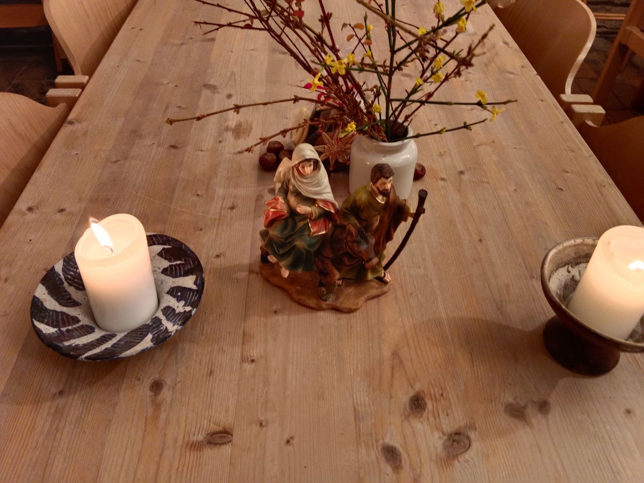 Ein Holztisch mit einer brennenden Kerze auf einem Teller, einer Keramikvase mit gelben Blumen und einer Figur von Maria und Josef mit einem Stab.
