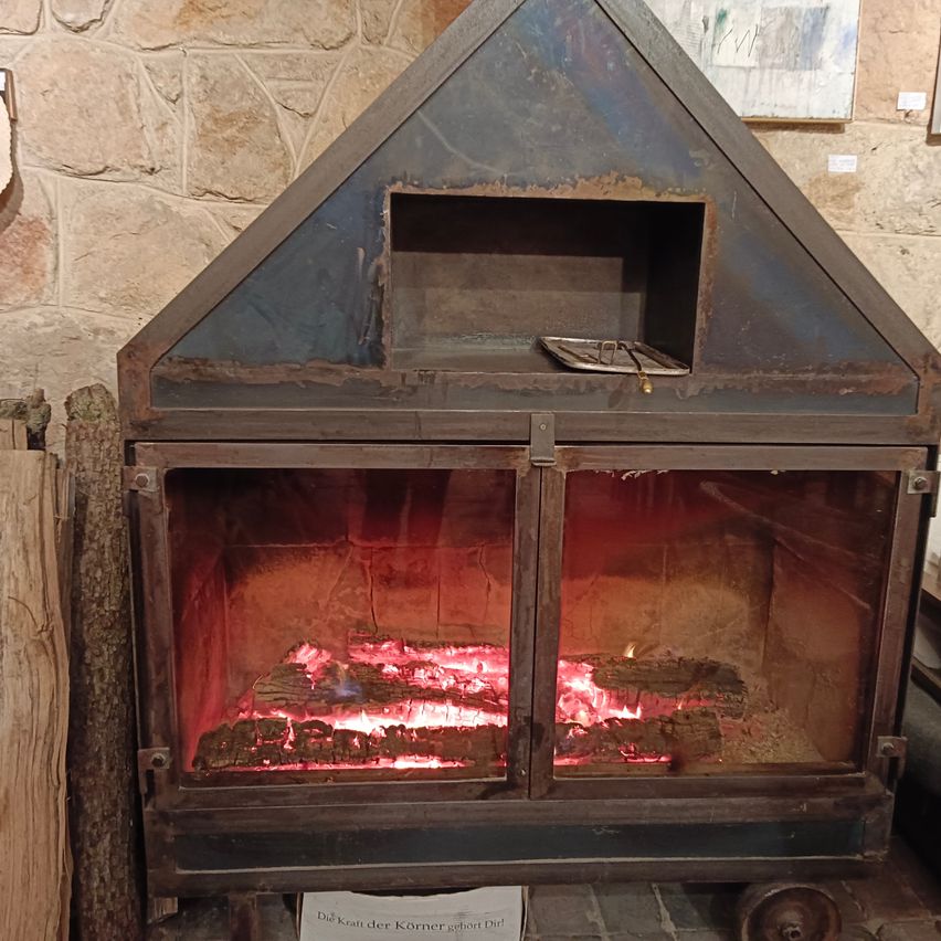 Ein alter Kamin mit Metalldeckel, offener Tür und einem brennenden Feuer im Inneren. Der Kamin steht auf einer Steinmauer.