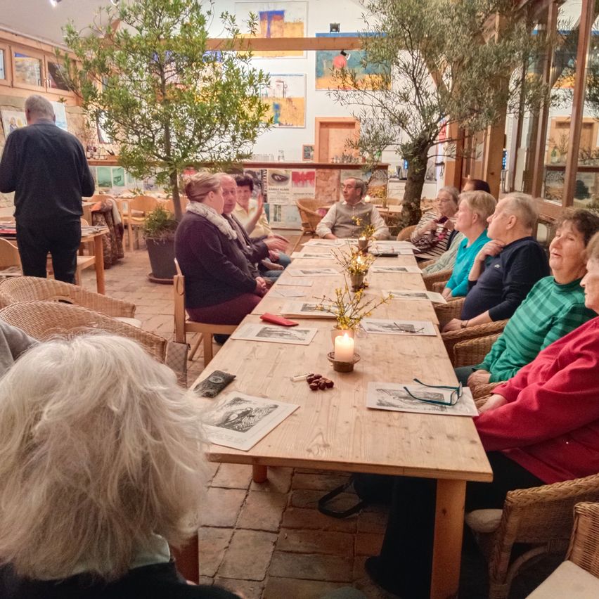 Eine Gruppe von Menschen sitzt um einen langen Holztisch in einem Raum, mit Bäumen und Kunstwerken an den Wänden. Ein Mann steht hinter ihnen und jeder hat Bücher und eine Kerze auf dem Tisch.