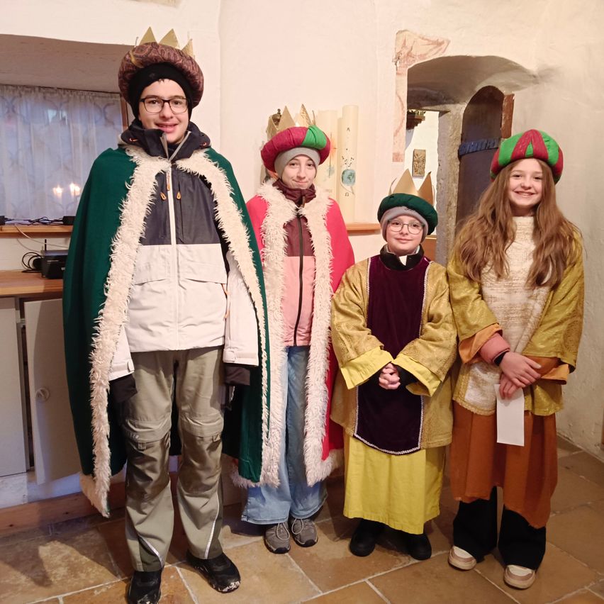 Fünf Personen in mittelalterlichen Kostümen posieren für ein Foto in einem Raum. Sie tragen alle Kronen und verschiedenfarbige Roben. Hinter ihnen ist eine Wand mit einem Fenster und einer Tür zu sehen.