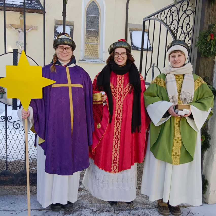 Drei Personen in liturgischen Gewändern und Mützen posieren im Winter vor einem Kircheneingang, eine hält einen Stern.