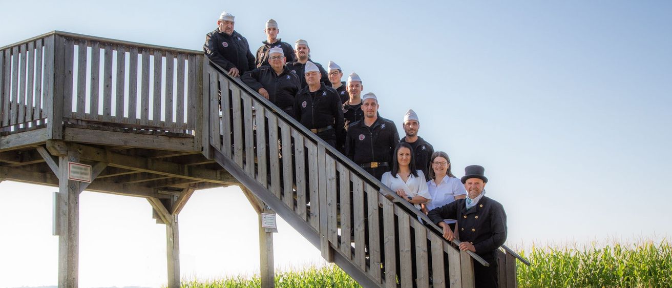 Eine Gruppe von Menschen in Uniformen, einige mit Mützen, steht auf einer Treppe. Zwei Frauen stehen unten, während andere auf den Stufen sind. Dahinter befindet sich ein Maisfeld.