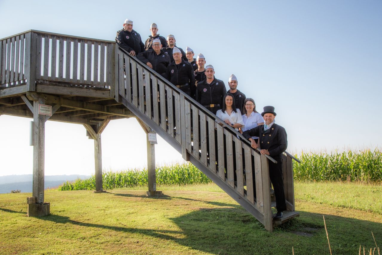 Eine Gruppe uniformierter Personen steht auf einer Holztreppe unter klarem Himmel, einige tragen Hüte. Ein Mann mit Topfhut steht unten. Die Umgebung ist grasig mit einem Maisfeld im Hintergrund.