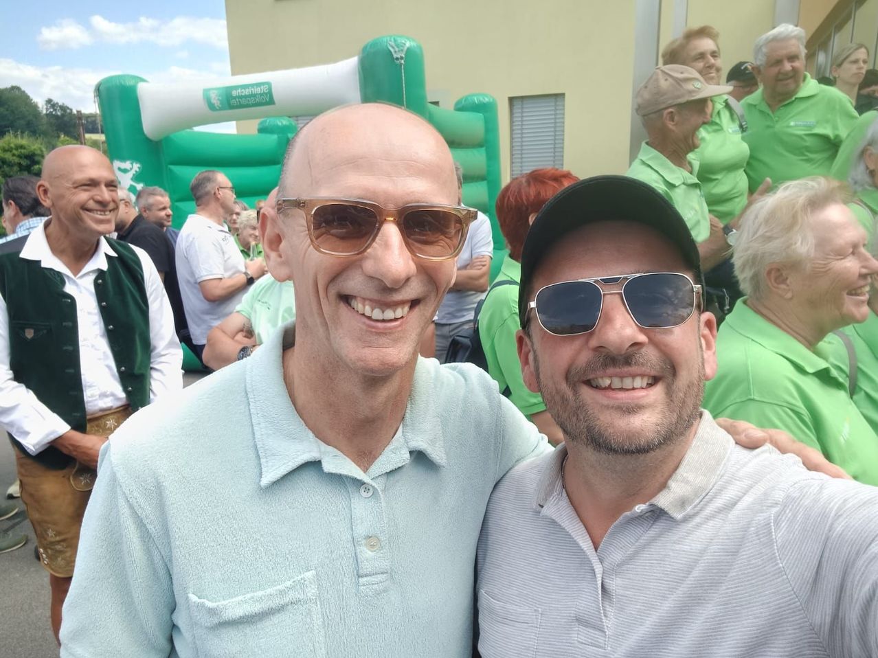 Zwei Männer lächeln und posieren für ein Foto vor einer Menschenmenge. Der Mann auf der linken Seite trägt ein hellblaues Poloshirt und eine Sonnenbrille. Der Mann auf der rechten Seite trägt ein gestreiftes Poloshirt und eine schwarze Kappe.