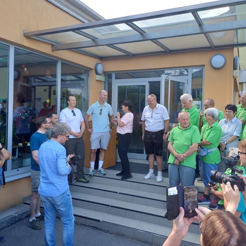 Eine Gruppe von Menschen in grünen Shirts und weißen Turnschuhen steht auf den Stufen eines Gebäudes mit Glastüren, während andere Fotos und Videos aufnehmen.